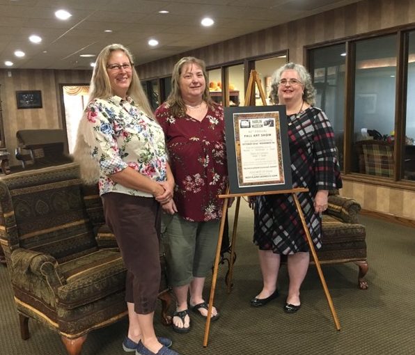 Three woman (left to right: Temi Lasater, Harlin Museum Board Treasurer; Pam Woodward, West Plains Savings & Loan Manager; Cleo Cockrum, Harlin Museum Board Vice President) stand side-by-side behind an easel that holds a poster announcing that West Plains Savings & Loan will donate $500 for the Harlin's 46th Annual Fall Art Show Best of Show award.